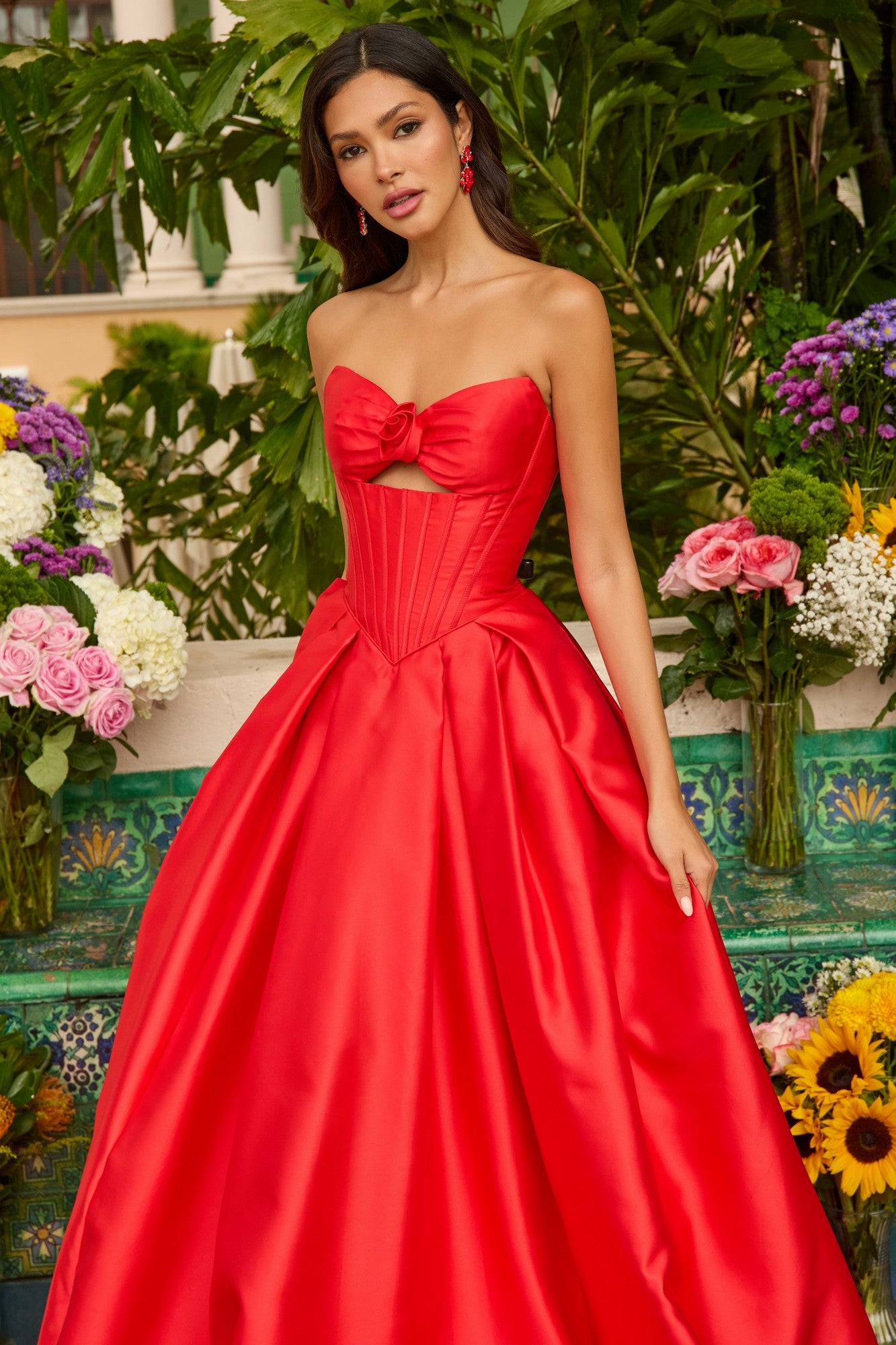 A woman stands before lush greenery and vibrant flowers, wearing the Ava Presley 47223 strapless satin prom ball gown in bright red with a bow detail on the bodice.